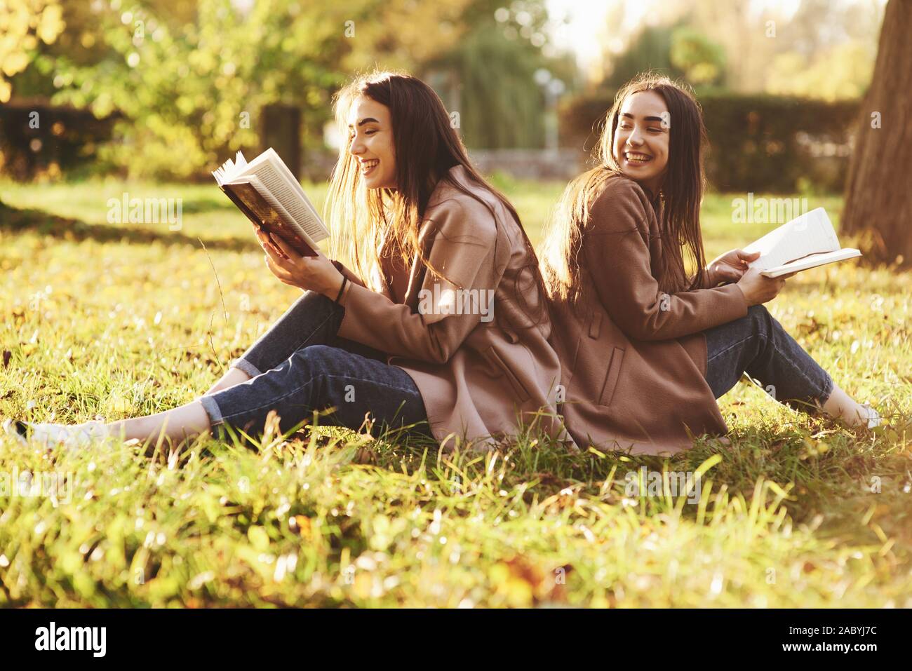 Laughinh brunette twin girls sitting back to back on the grass and having fun with legs slightly bent in knees, with brown books in hands, wearing Stock Photo