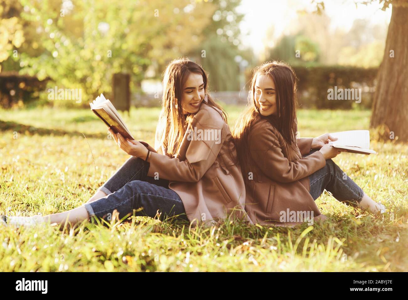 Smiling brunette twin girls sitting back to back on the grass and looking at each other, legs slightly bent in knees, with brown books in hands Stock Photo
