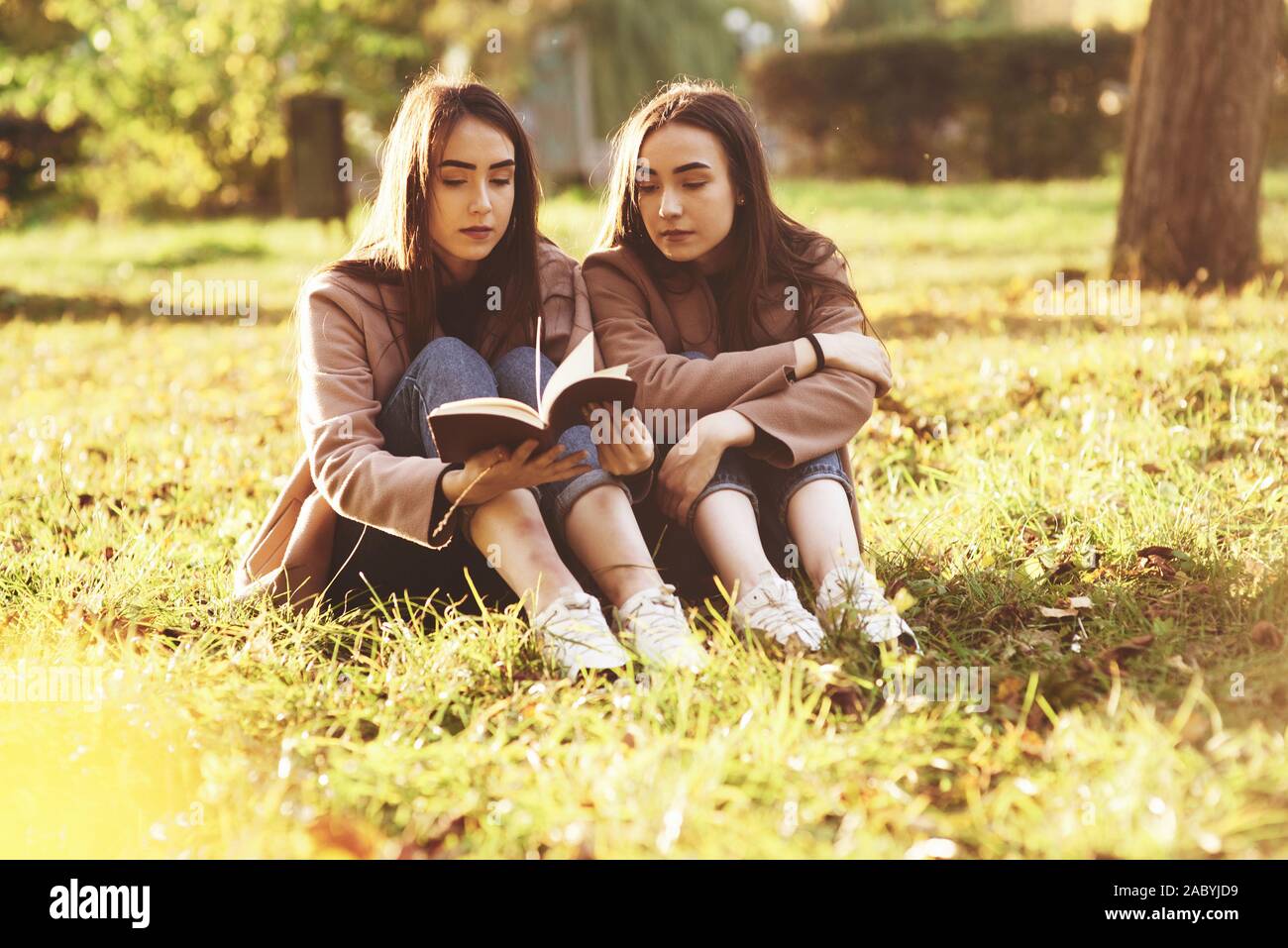 Young pretty brunette twin girls sitting on the grass with legs slightly bent in knees and reading in a brown book, wearing casual coat in autumn Stock Photo