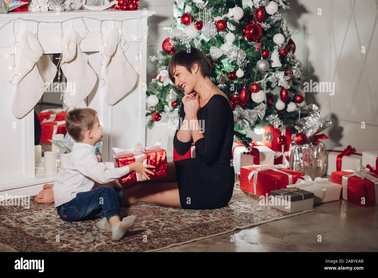 https://c8.alamy.com/comp/2ABYKAB/son-gives-a-christmas-gift-to-mom-mom-pleasantly-surprised-and-cute-looks-at-the-child-isolated-on-a-new-year-tree-background-with-gifts-a-fireplac-2ABYKAB.jpg