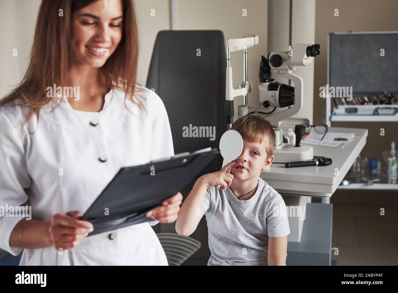 Woman smiling during the process. Doing testing in the oculist office with child. Doctor notes data Stock Photo