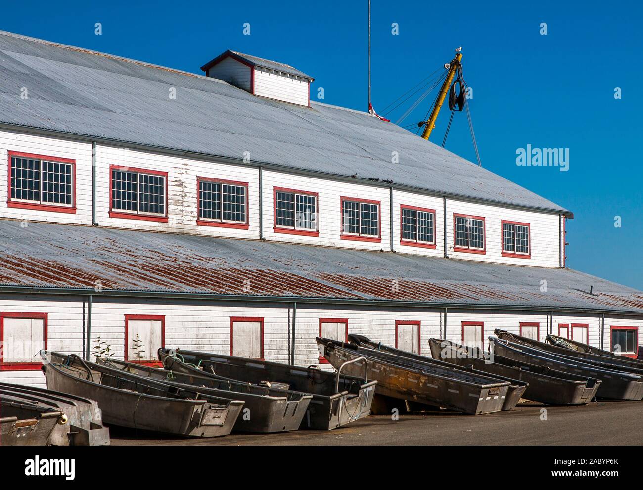 Weathered metal fishing boats, all of the same type and shape, out of water, lined up side by side along the wall of old fishing warehouse, rusty roof Stock Photo