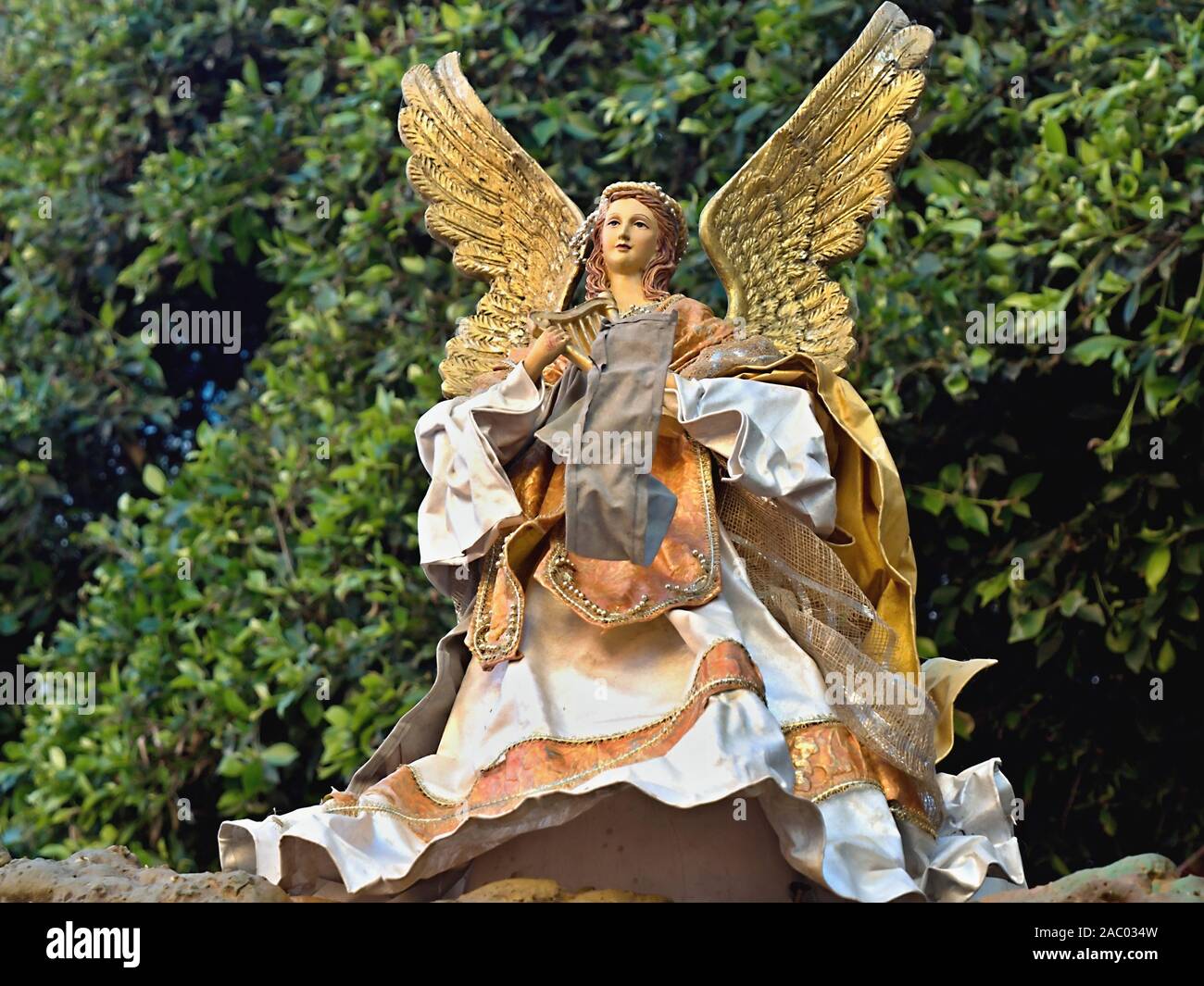big Christmas angel with golden wings and lush robe in gold and beige. he stands in front of an evergreen bush.The angel has a harp in her hand is fem Stock Photo