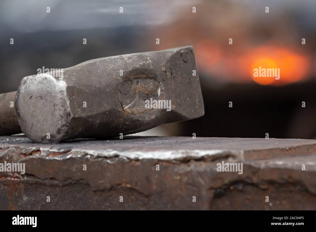 Old rusty hammer lies on the anvil with flame of brazier forge on background. Blacksmith, metalsmith, farrier tools. Close view. Stock Photo