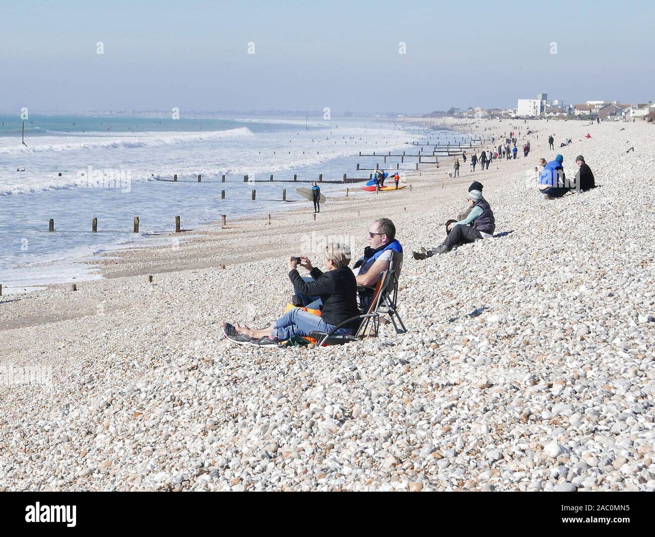 Winter surfing at Selsey beach in February 2019 with people sitting on the beach watching, sea groins in the background Stock Photo