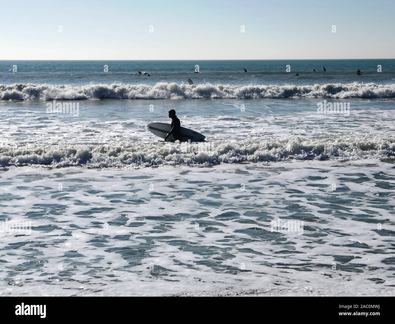 Winter surfing at Selsey beach in February 2019 with a surfer walking in the waves holding his surfboard Stock Photo
