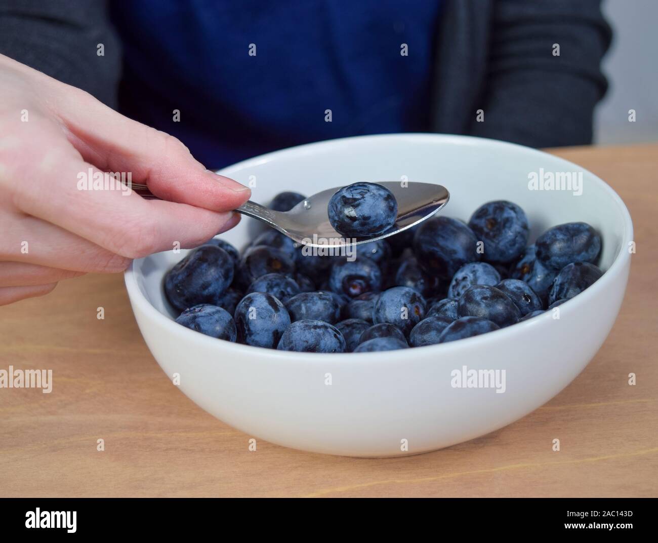 Closeup of Caucasian woman dipping a spoon into a bowl of fresh organic blueberries. The bowl of blueberries rests on a rustic wooden table. Stock Photo