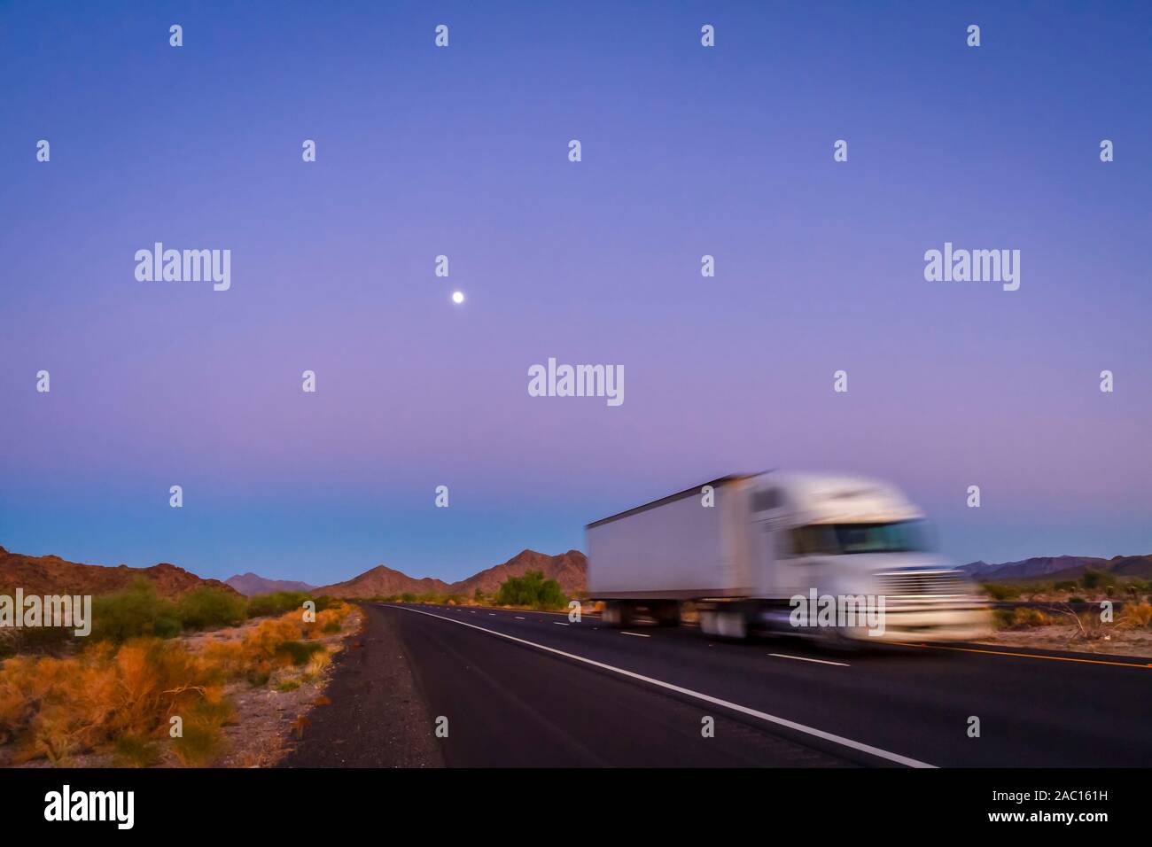 Deliberately blurred 18 wheel long haul truck on highway in desert at purple and blue sunrise with moon in the sky Stock Photo