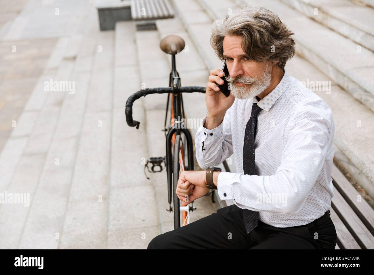 Photo of handsome mature businessman in formal clothes talking on cellphone while sitting on bench with bicycle outdoors Stock Photo