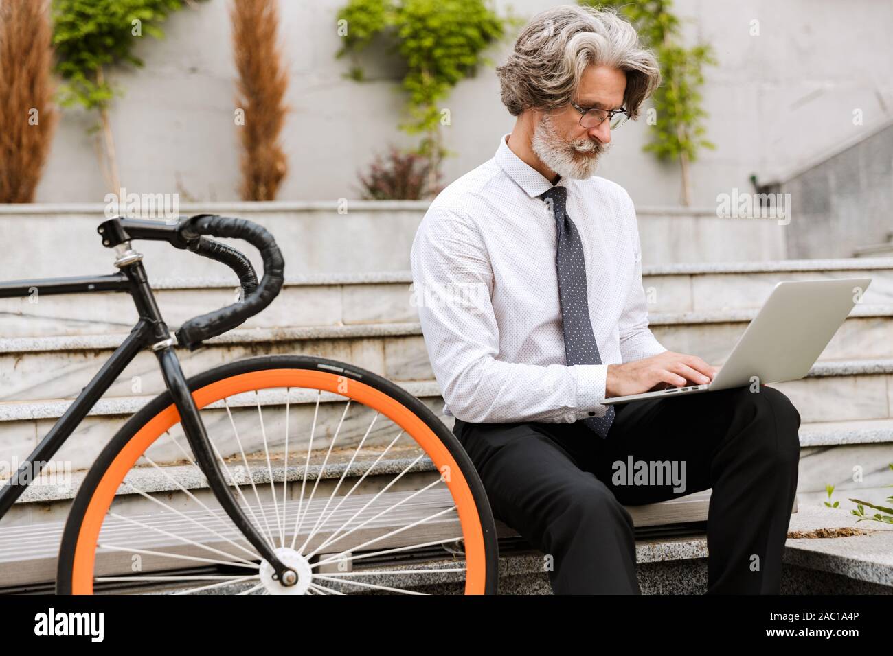 Photo of handsome mature businessman in eyeglasses typing on laptop while sitting on bench with bicycle outdoors Stock Photo