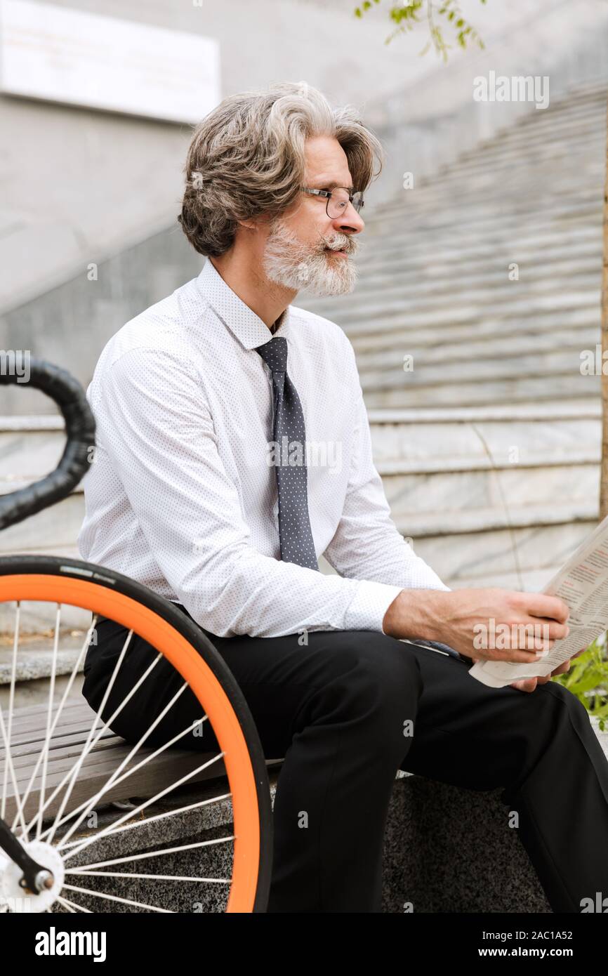 Photo of gray-haired confident businessman in eyeglasses reading newspaper while sitting on bench with bicycle outdoors Stock Photo