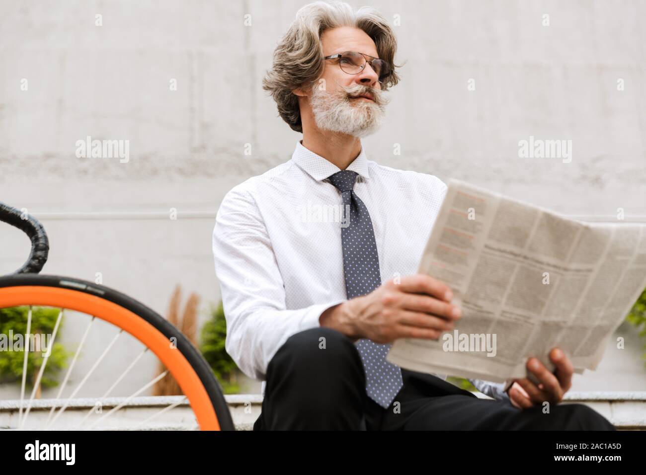 Photo of gray-haired bearded businessman in eyeglasses reading newspaper while sitting on bench with bicycle outdoors Stock Photo