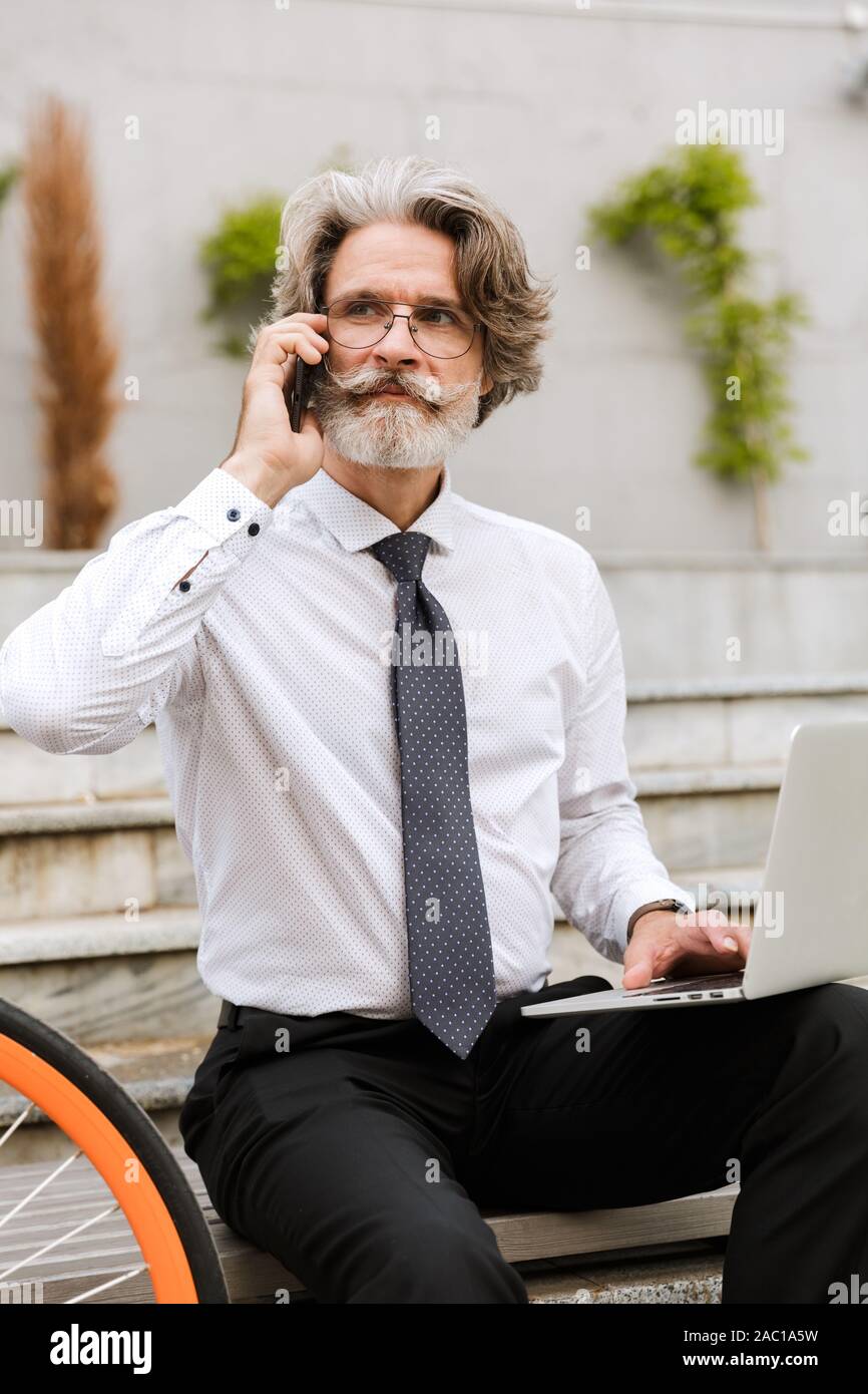 Photo of mature confident businessman in eyeglasses using cellphone and laptop while sitting on bench with bicycle outdoors Stock Photo