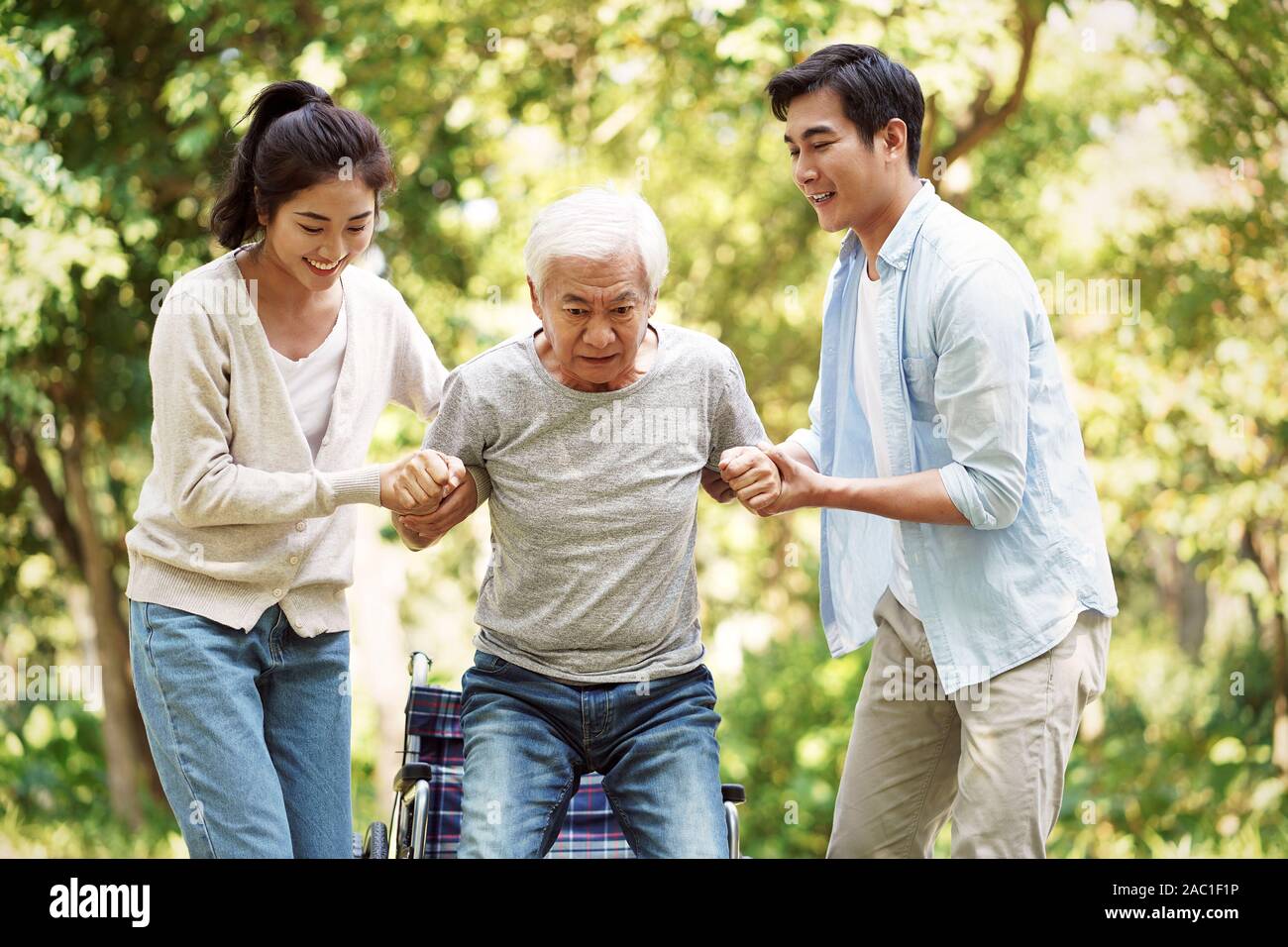 young asian man and woman helping wheelchair bound senior man stand up and walk Stock Photo
