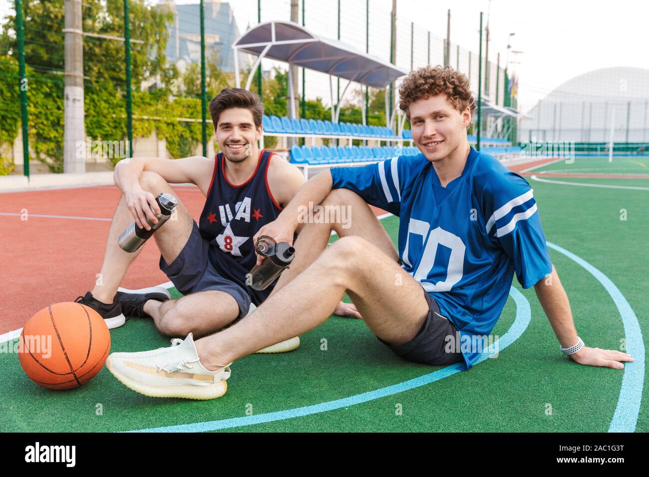 Two attarctive fit men basketball players sitting on a sports ground, looking at camera Stock Photo