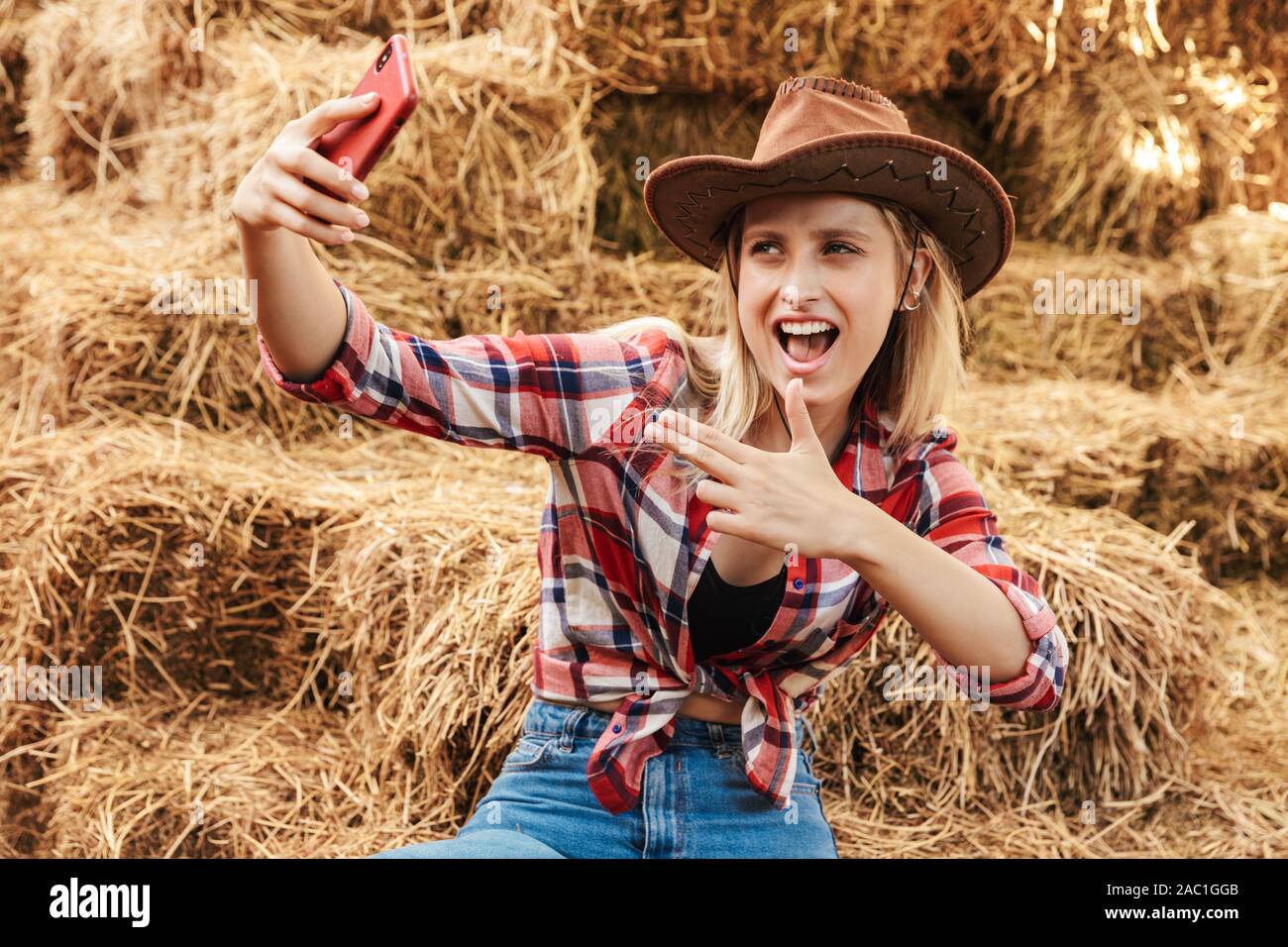 Smiling cheerful young blonde cowgirl sitting on a haystack at the barn, taking a selfie Stock Photo