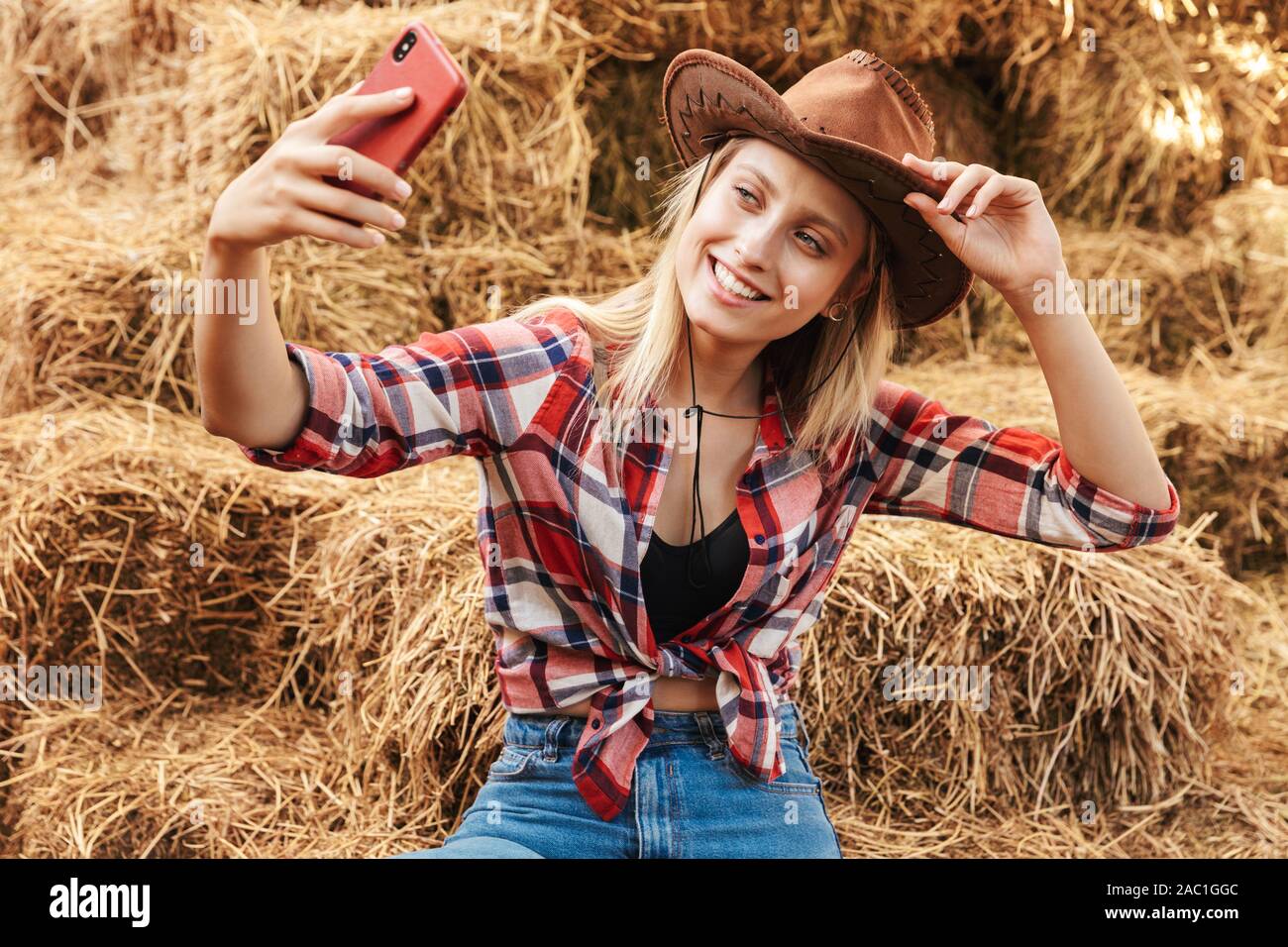 Smiling cheerful young blonde cowgirl sitting on a haystack at the barn, taking a selfie Stock Photo