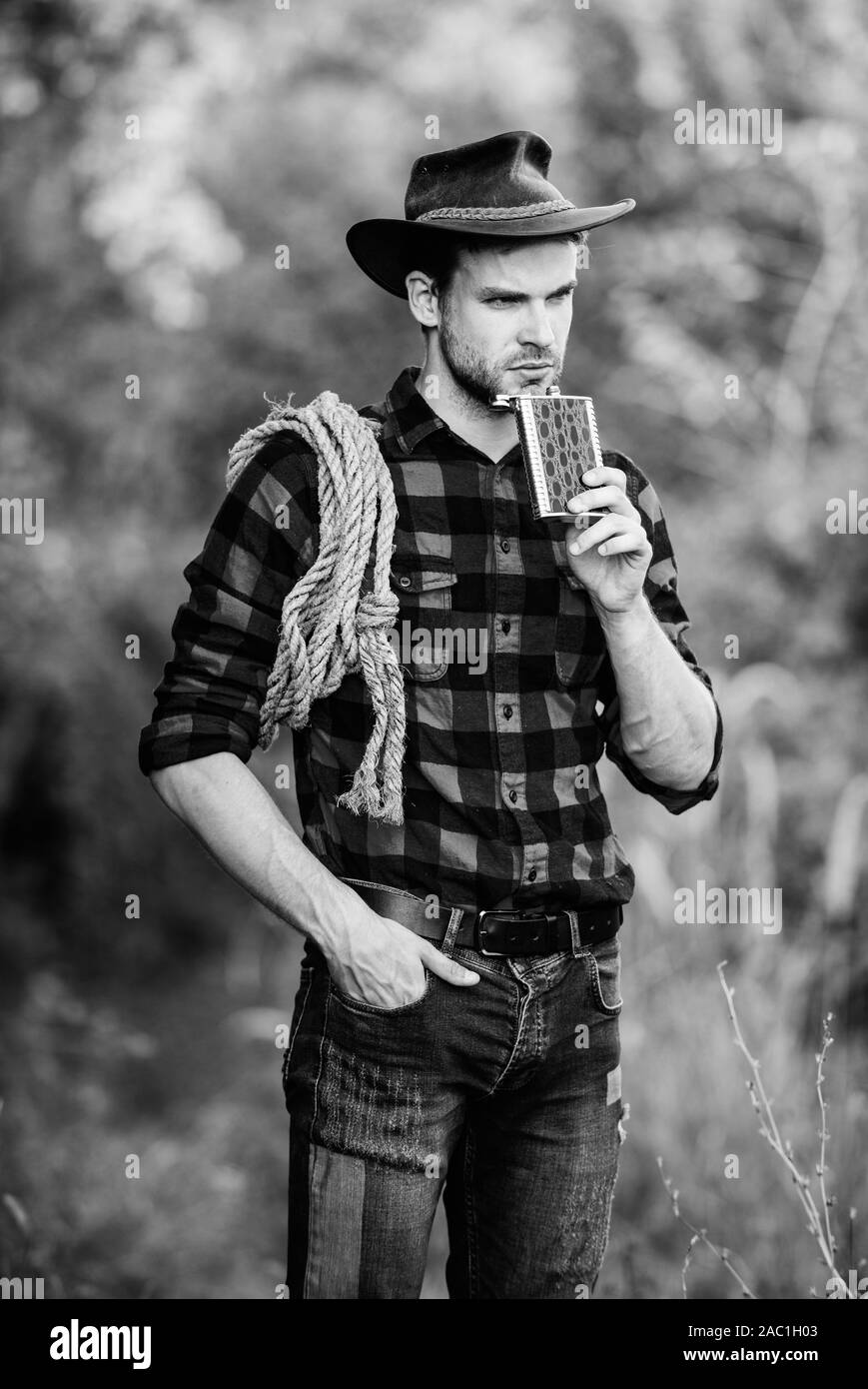 Cowboy ranch worker. Brutal cowboy drinking alcohol. Man handsome cowboy nature background. Bourbon whiskey. Western culture. Man wearing hat hold rope and flask. Lasso tool American cowboy. Stock Photo