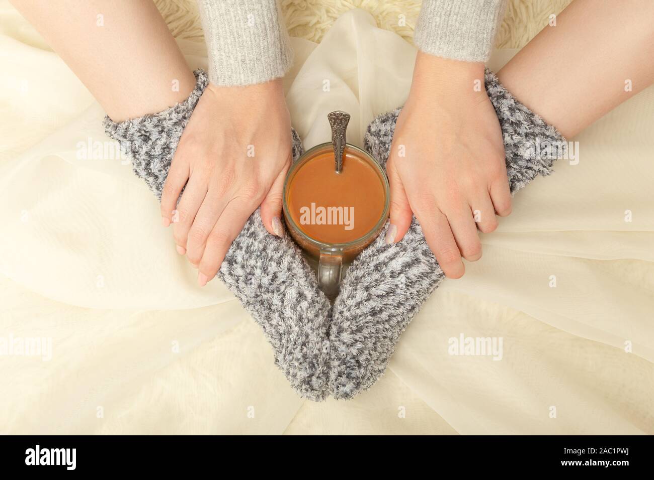unrecognizable young woman in sweater and woolen socks with cup of hot tea, closeup hands and feet, monochrome Stock Photo