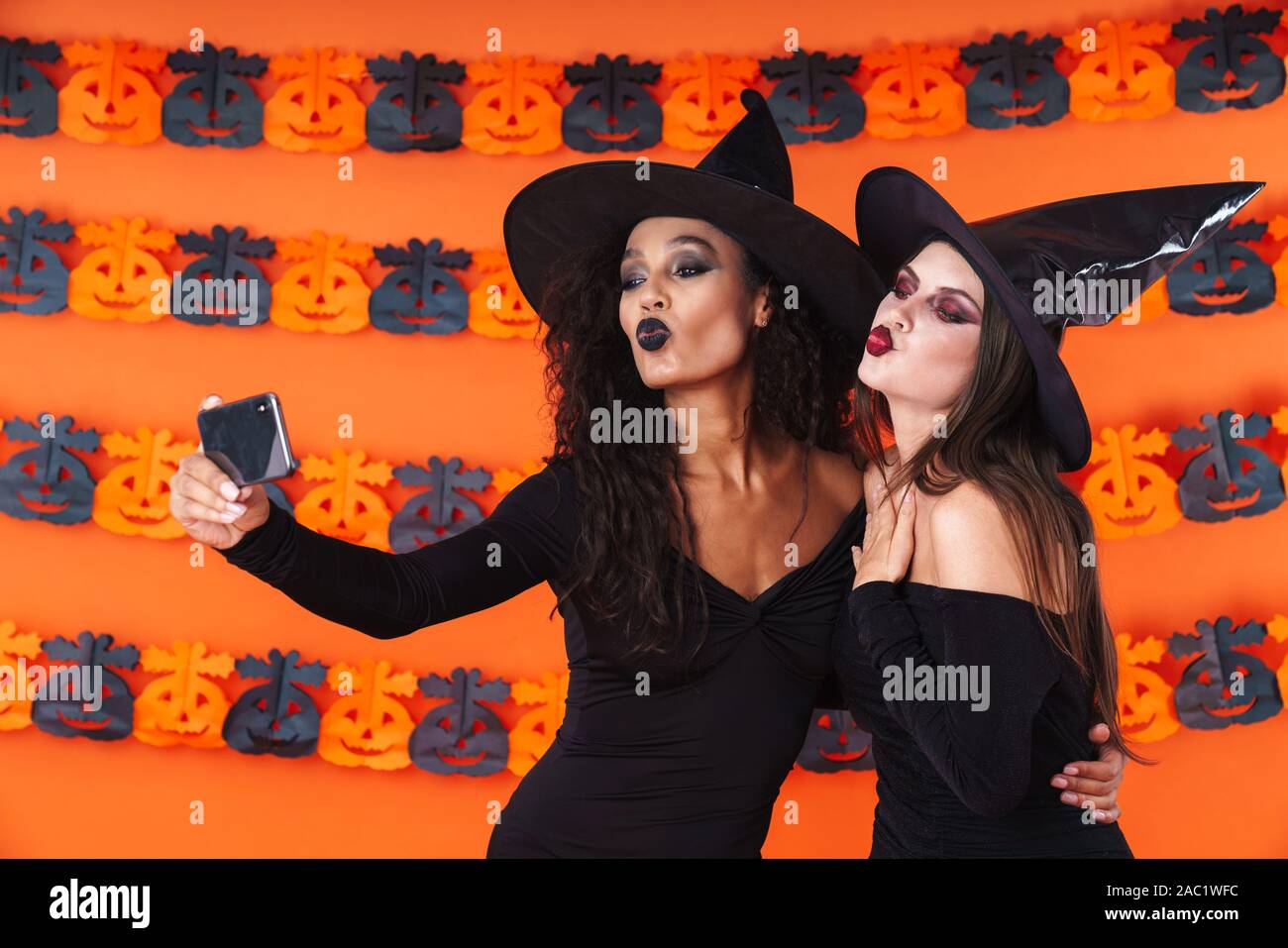 Image of cute witch women in black halloween costume taking selfie photo on cellphone with kissing isolated over orange pumpkin wall Stock Photo