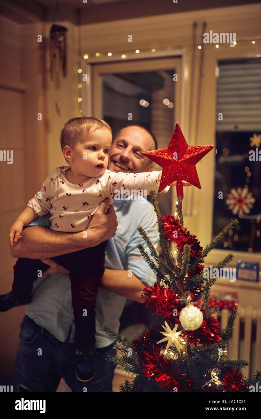 baby and smiling father putting a star on the top of the Christmas tree Stock Photo
