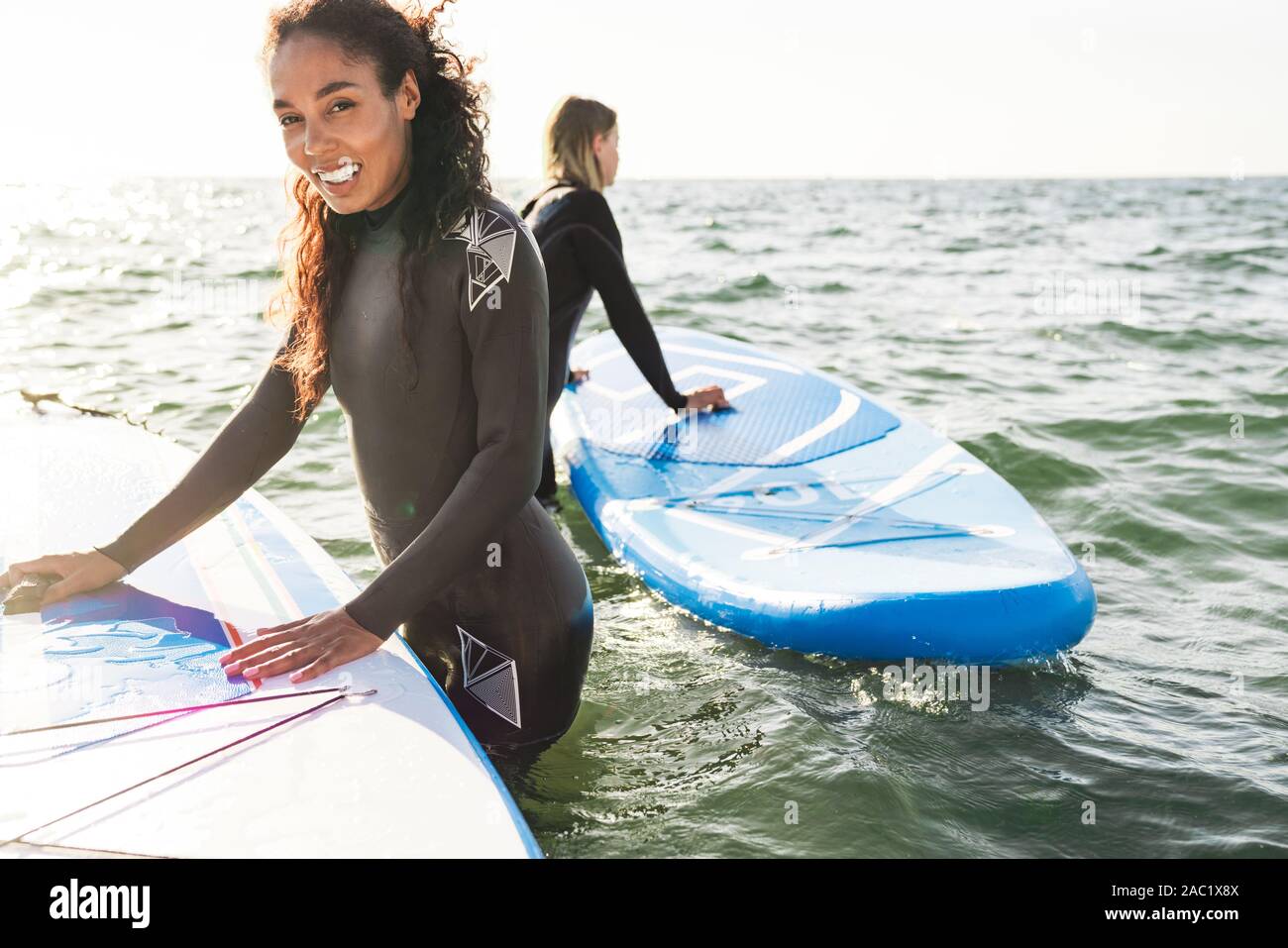 Image of happy multinational women in wetsuits using surfboard and laughing while working out on water Stock Photo