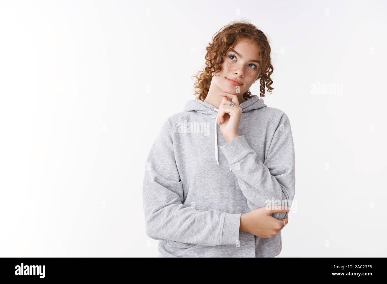 Thoughtful cute dreamy young caucasian ginger girl with freckles pimples wearing grey hoodie thinking look doubtful up make decision remember number Stock Photo