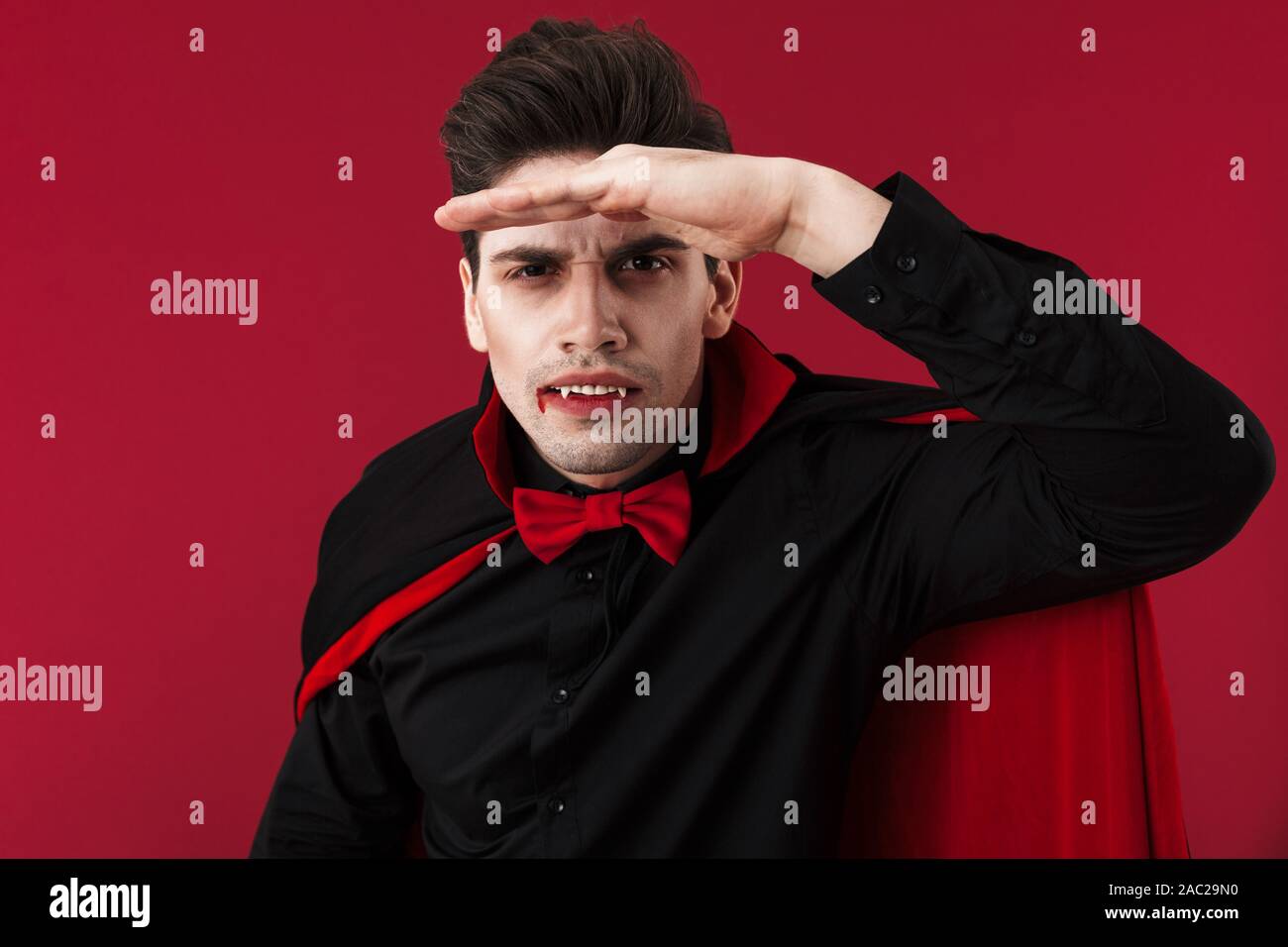 Image of handsome vampire man with blood and fangs in black halloween costume looking at camera isolated over red wall Stock Photo