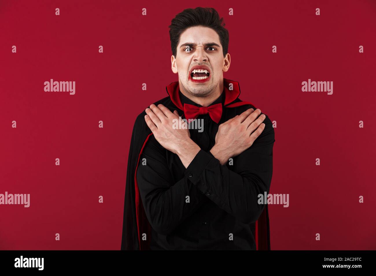 Image of frightening vampire man with blood and fangs in black halloween costume standing as undead isolated over red wall Stock Photo