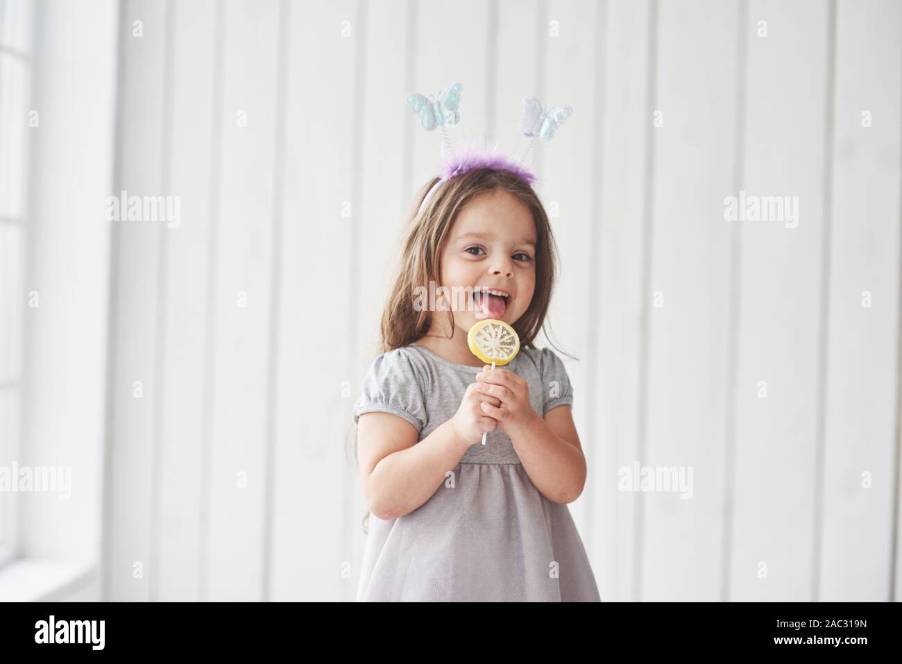 Smiling and feeling joy. Pretty little girl eating the yellow candy with toy butterflies on his head in the white room Stock Photo
