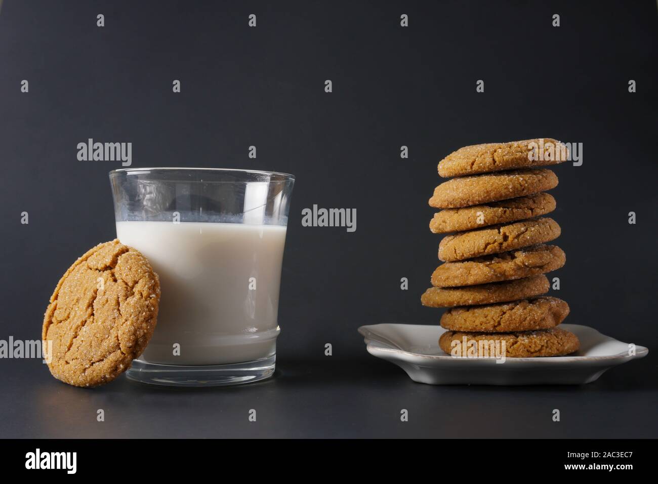 A ginger cookie rests on a glass of milk next to a stack of ginger cookies on a white plate and a black background; copy space Stock Photo