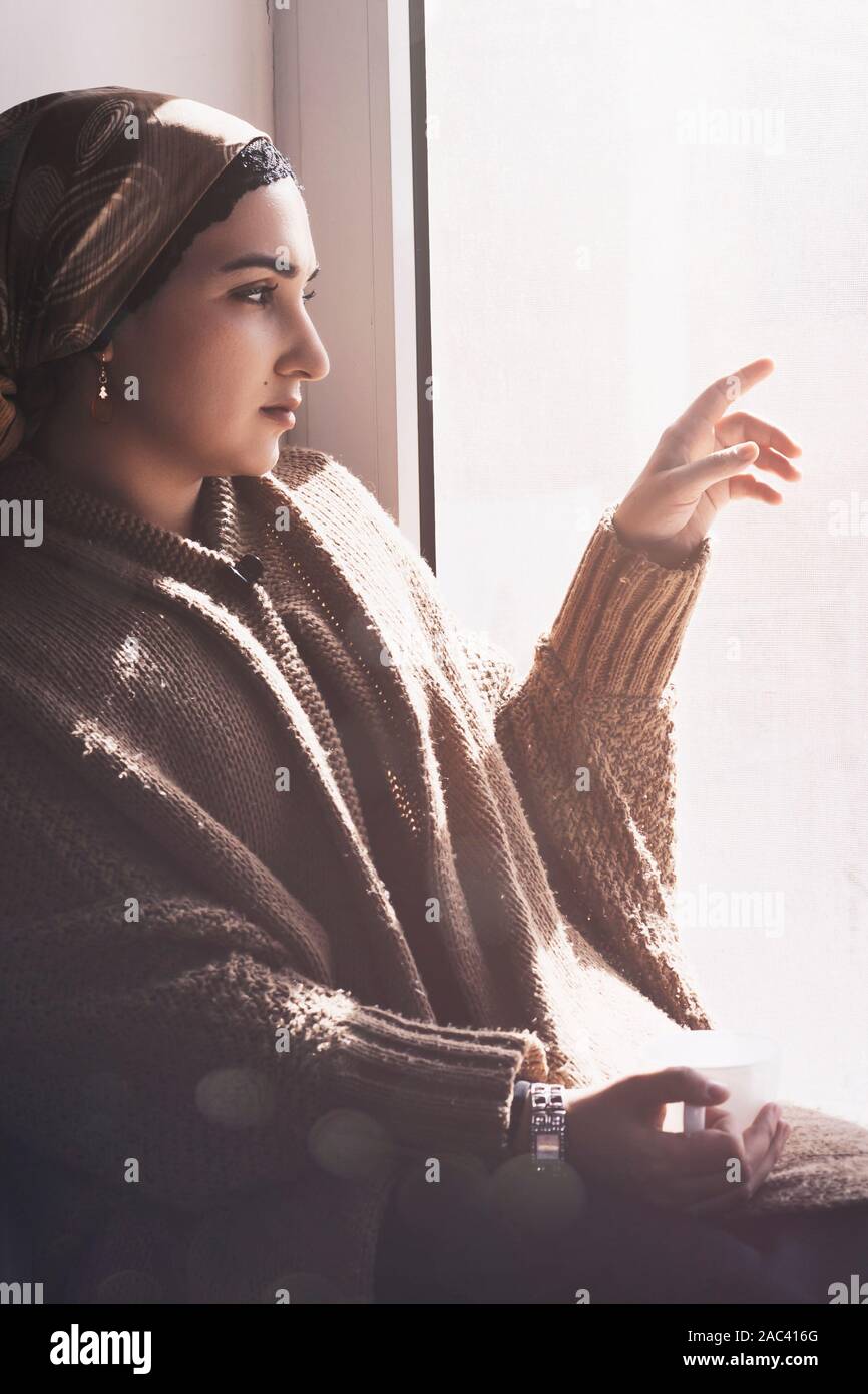 Young Muslim woman sits on window sill and looking away. Depressed middle-eastern female near the window at home. Lonely cheerless girl feeling stress Stock Photo