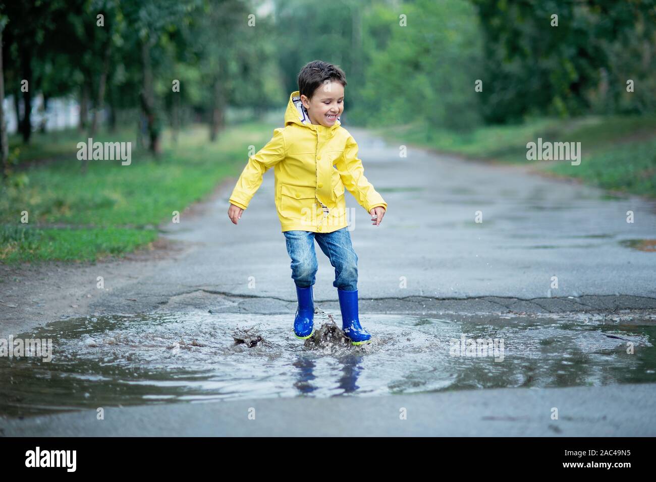 A wet child is jumping in a puddle. Fun on the street. Tempering in summer. Splashes, drops of water, outdoor. waterproof boots jump in puddle and mud Stock Photo