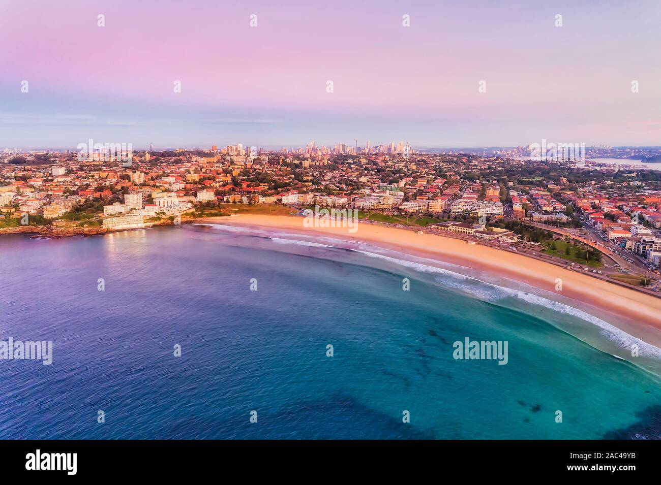 Smooth waters of Pacific ocean surfing on famous Sydney Bondi beach at sunrise in aerial view towards distant city CBD towers and Sydney harbour bridg Stock Photo