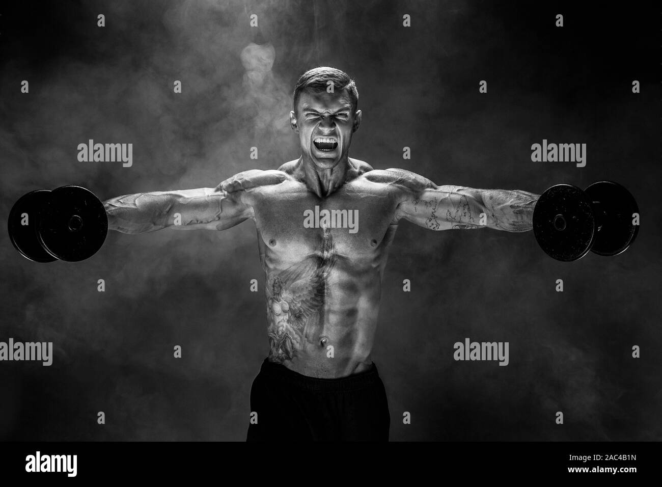 very brawny guy bodybuilder, execute exercise with dumbbells, on deltoid muscle shoulder. Sream for motivation. Shot on studio smoke background. Stock Photo