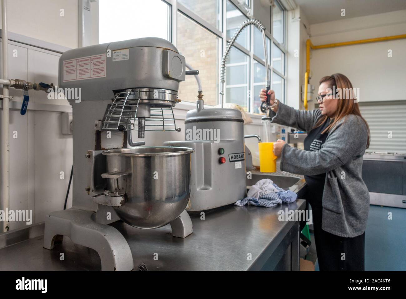A lady gets water in a jug from a tap in a canteen kitchen. Equipment including an industrial food processor sit on a stainless steel worktop. Stock Photo