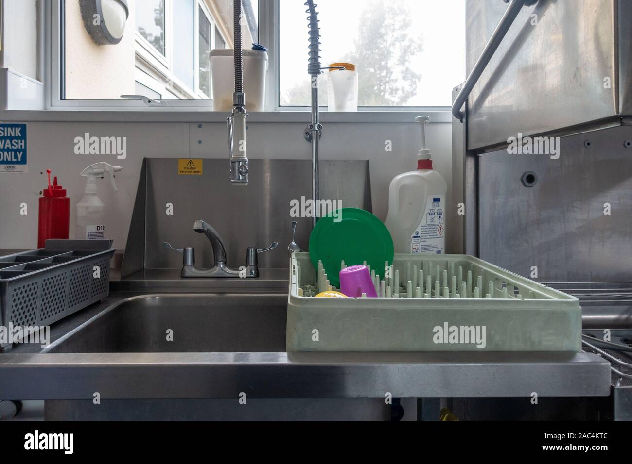A tray from an industrial dishwasher sits on the side of a sink in a school kitchen. Stock Photo