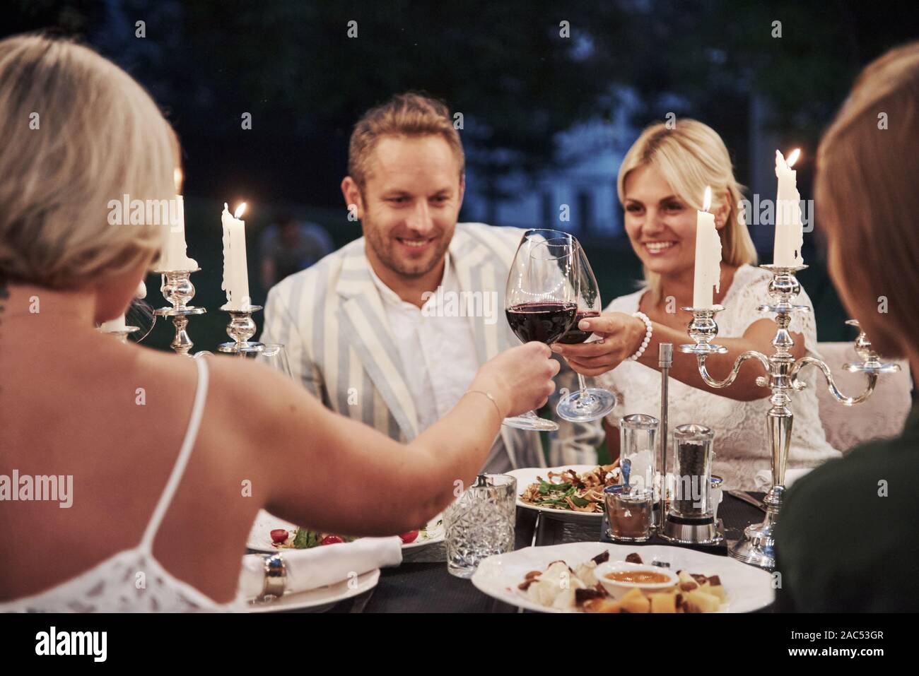 Knocking the glasses with wine. Group of friends in the elegant wear have luxury dinner Stock Photo