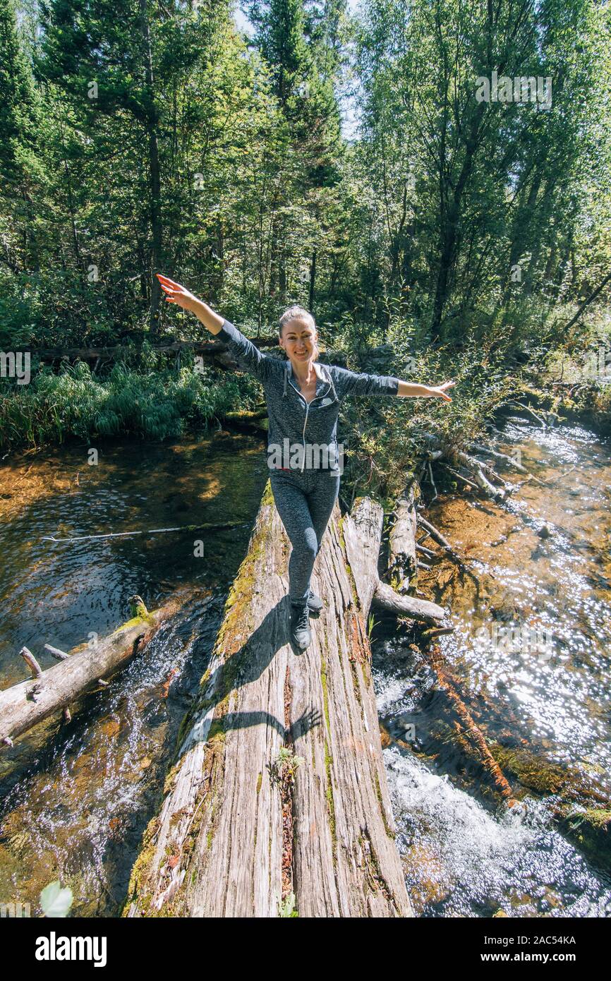 Beautiful girl stand on log and chilling after travel. Woman traveler in summer passes on wooden bridge in background of forest. Bridged lies on the r Stock Photo