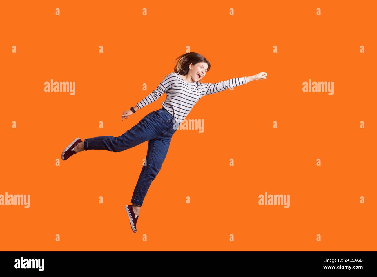 Superhero. Portrait of joyous pretty woman with brown hair in long sleeve shirt jumping high with one stretched arm, feeling to be superman flying up. Stock Photo