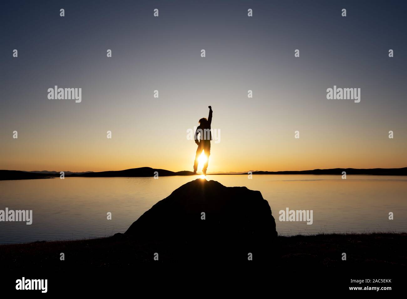 Man's silhouette jumps on big rock against sunset lake and mountains Stock Photo