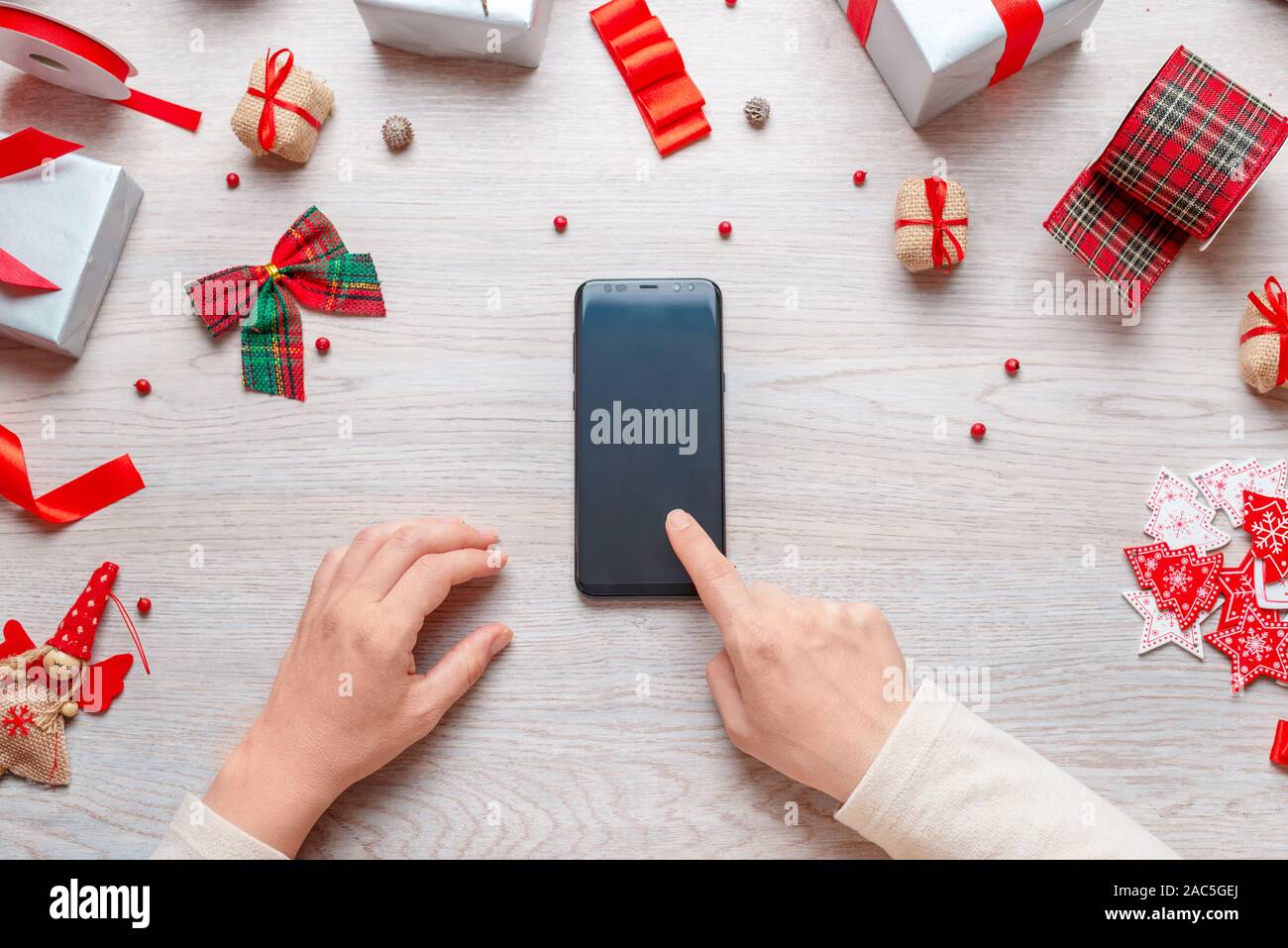 Hand touch blank phone display on a wooden desk surrounded by Christmas decorations and gifts. Concept of online shopping at Christmas time. Top view Stock Photo