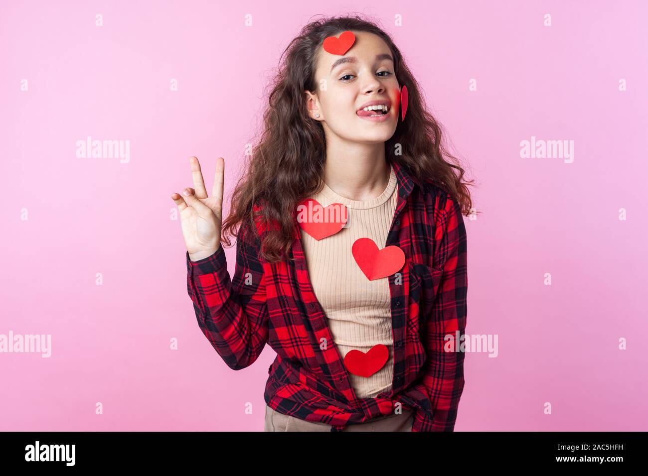Valentines Day. Portrait of playful cute teen girl with curly brunette hair and paper heart stickers on her face and clothes showing tongue and gestur Stock Photo