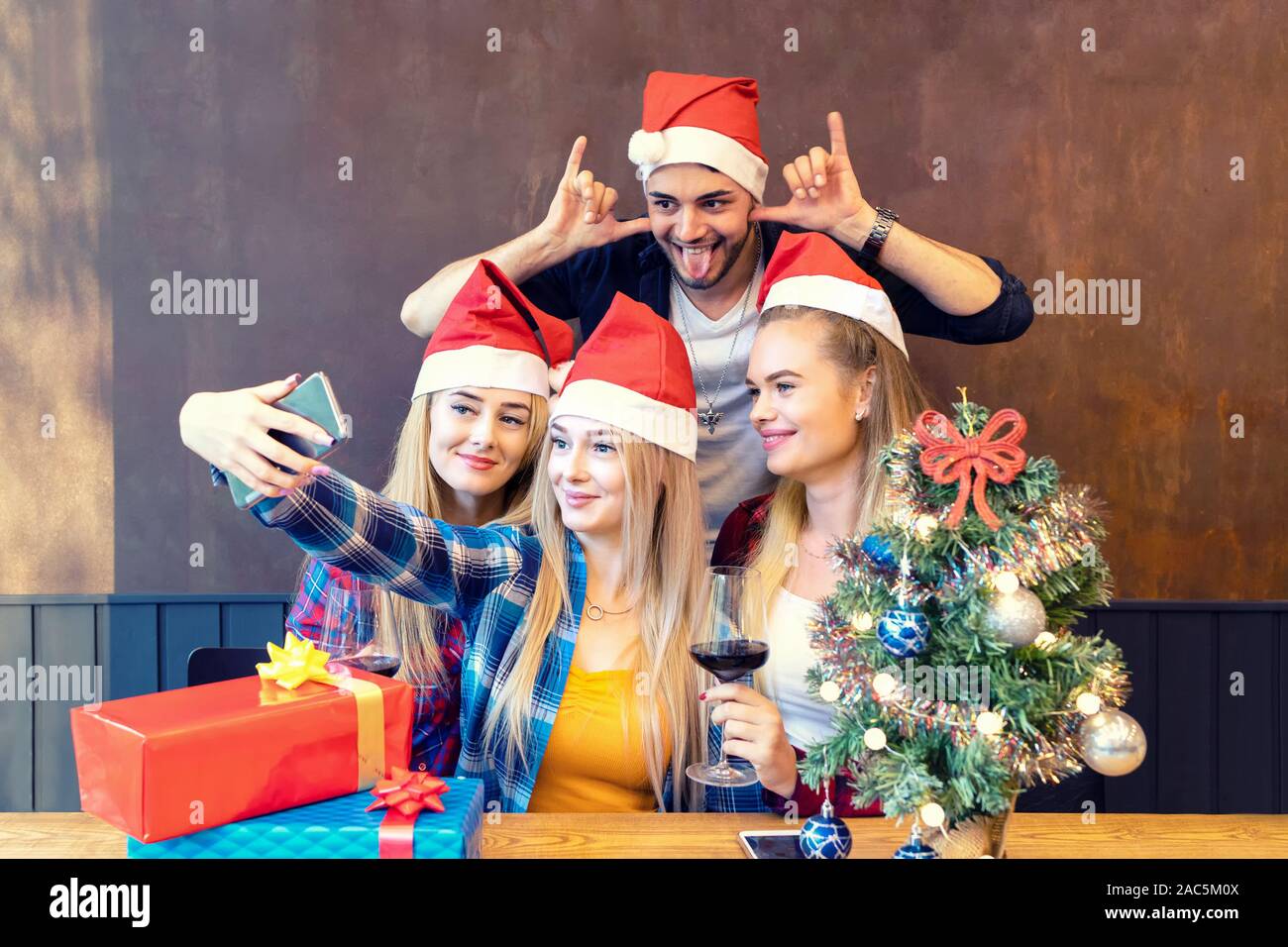 Happy young friends wearing Santa claus hat enjoying Christmas party taking selfie with funny faces at home sharing it on social media Stock Photo