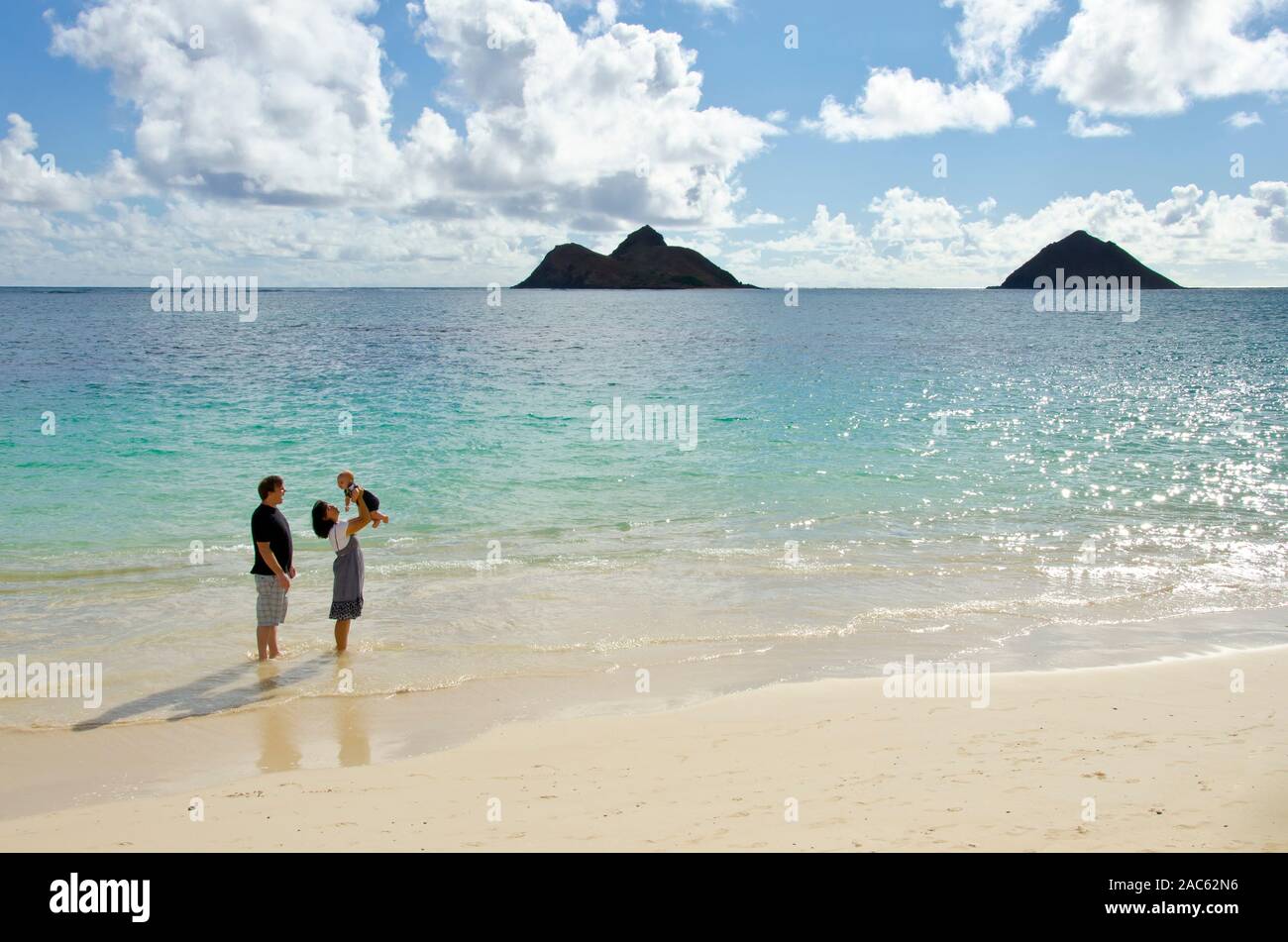 A father looking on while a mother lifts their son on Lanikai Beach, O'ahu, with the Mokulua Islands in the distance. Stock Photo