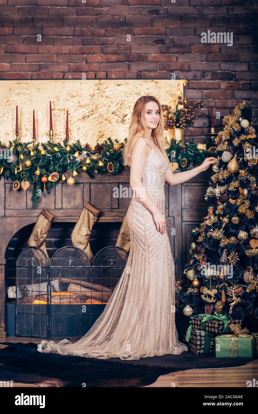 Beautiful young blonde girl in a long Golden dress smiling and decorating  the Christmas tree near the fireplace and the brick wall. New year, holiday  Stock Photo - Alamy