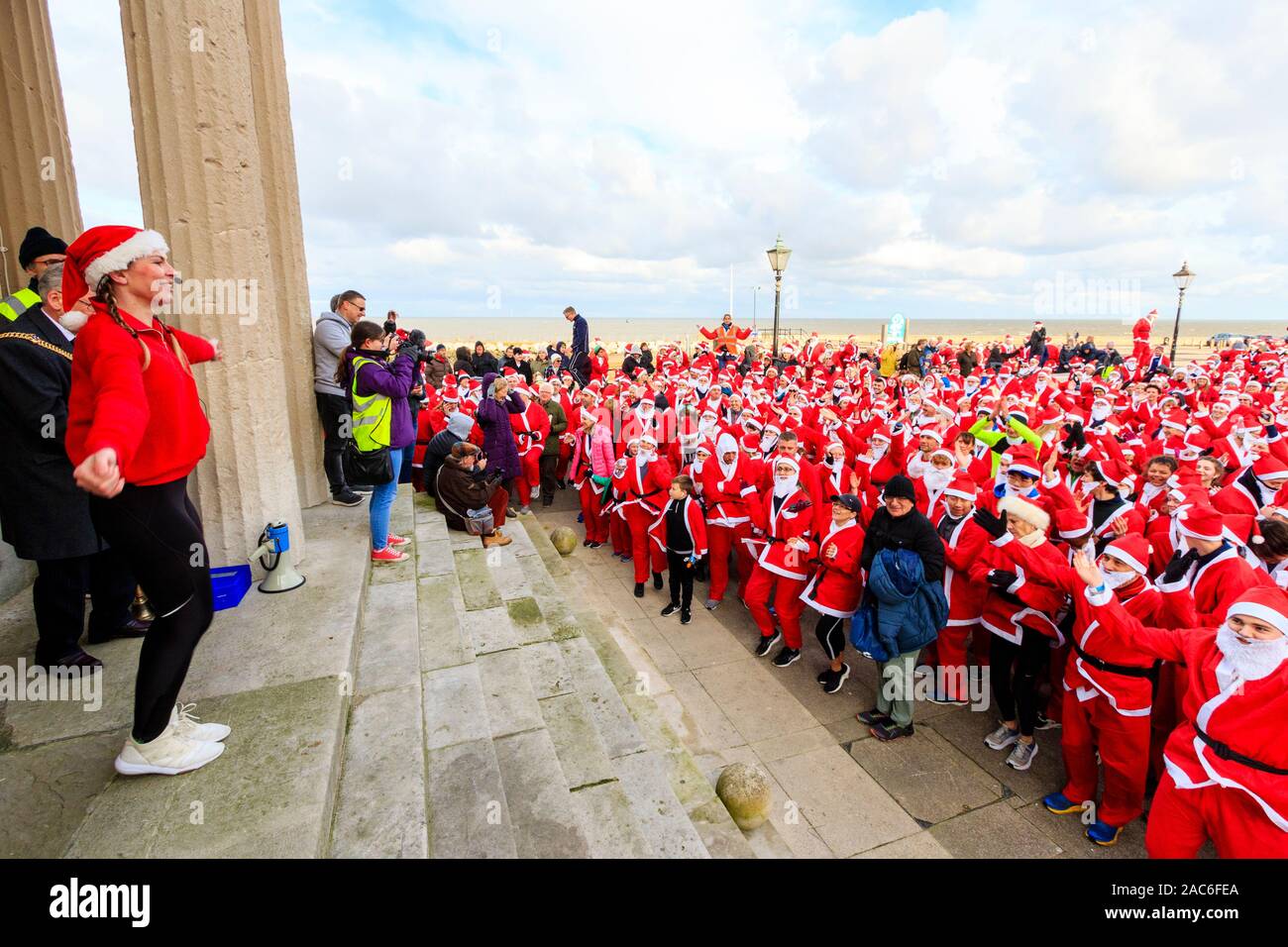 Young woman fitness trainer standing on top of steps directing group exercise with a crowd of people all dressed up as Father Christmas at the bottom of the steps on the seafront at Herne Bay in Kent, England. 'Santa's on the Run' Pilgrims hospices annual charity event. Stock Photo