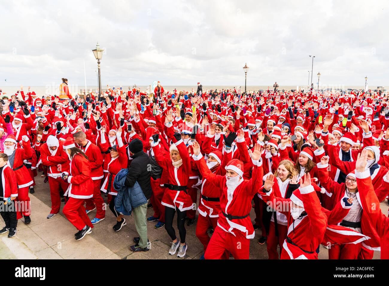 High angle view of a crowd of people all dressed up as Father Christmas on the seafront at Herne Bay in Kent, England. Most have arms raised as they follow instruction during a group exercise just before the charity event , 'Santa on the run' for the Pilgrims hospices. Stock Photo