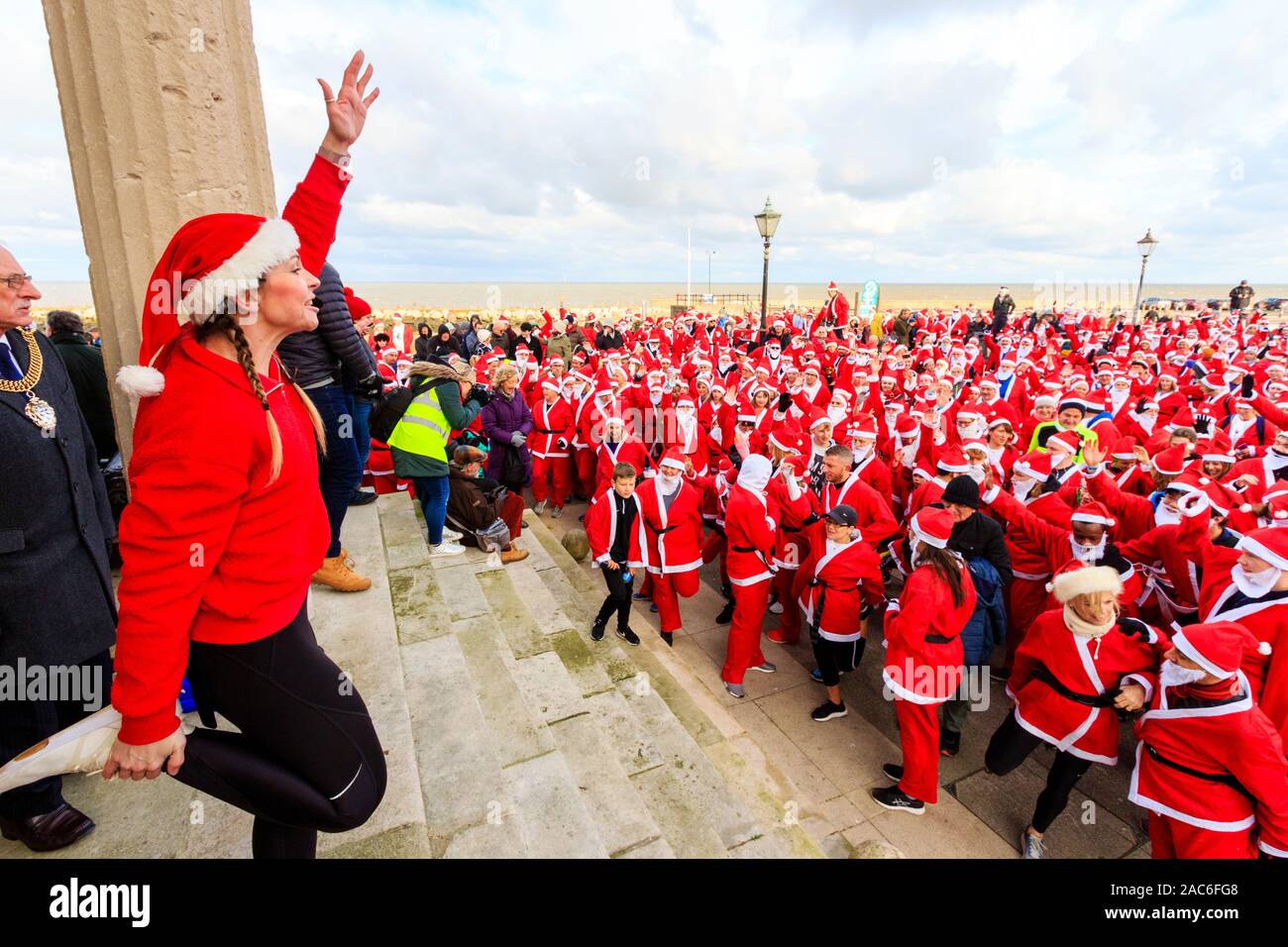 Young woman fitness trainer standing on top of steps directing group exercise with a crowd of people all dressed up as Father Christmas at the bottom of the steps on the seafront at Herne Bay in Kent, England. 'Santa's on the Run' Pilgrims hospices annual charity event. Stock Photo