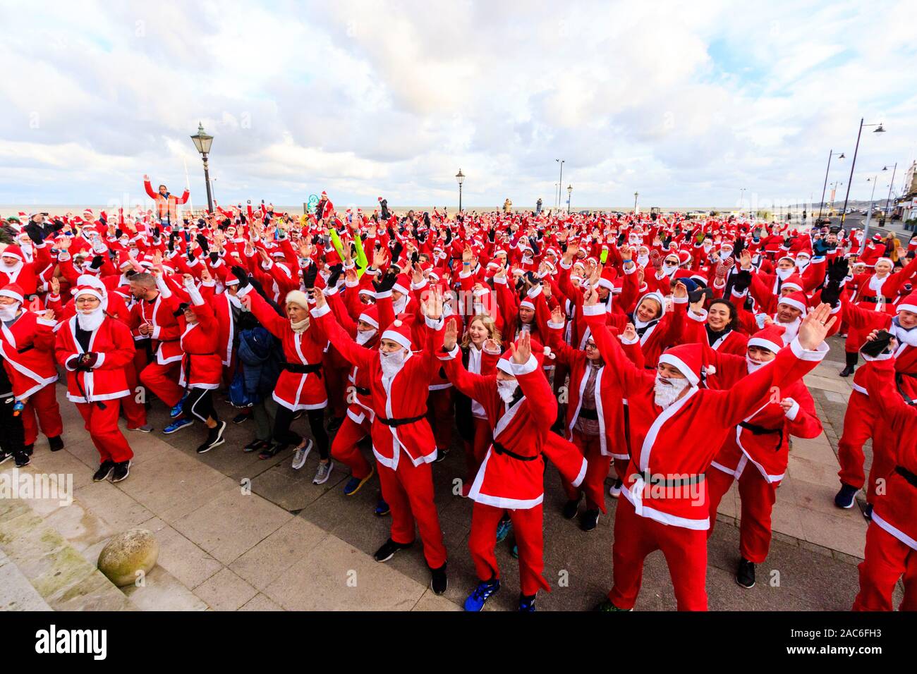 High angle view of a crowd of people all dressed up as Father Christmas on the seafront at Herne Bay in Kent, England. Most have arms raised as they follow instruction during a group exercise just before the charity event , 'Santa on the run' for the Pilgrims hospices. Stock Photo